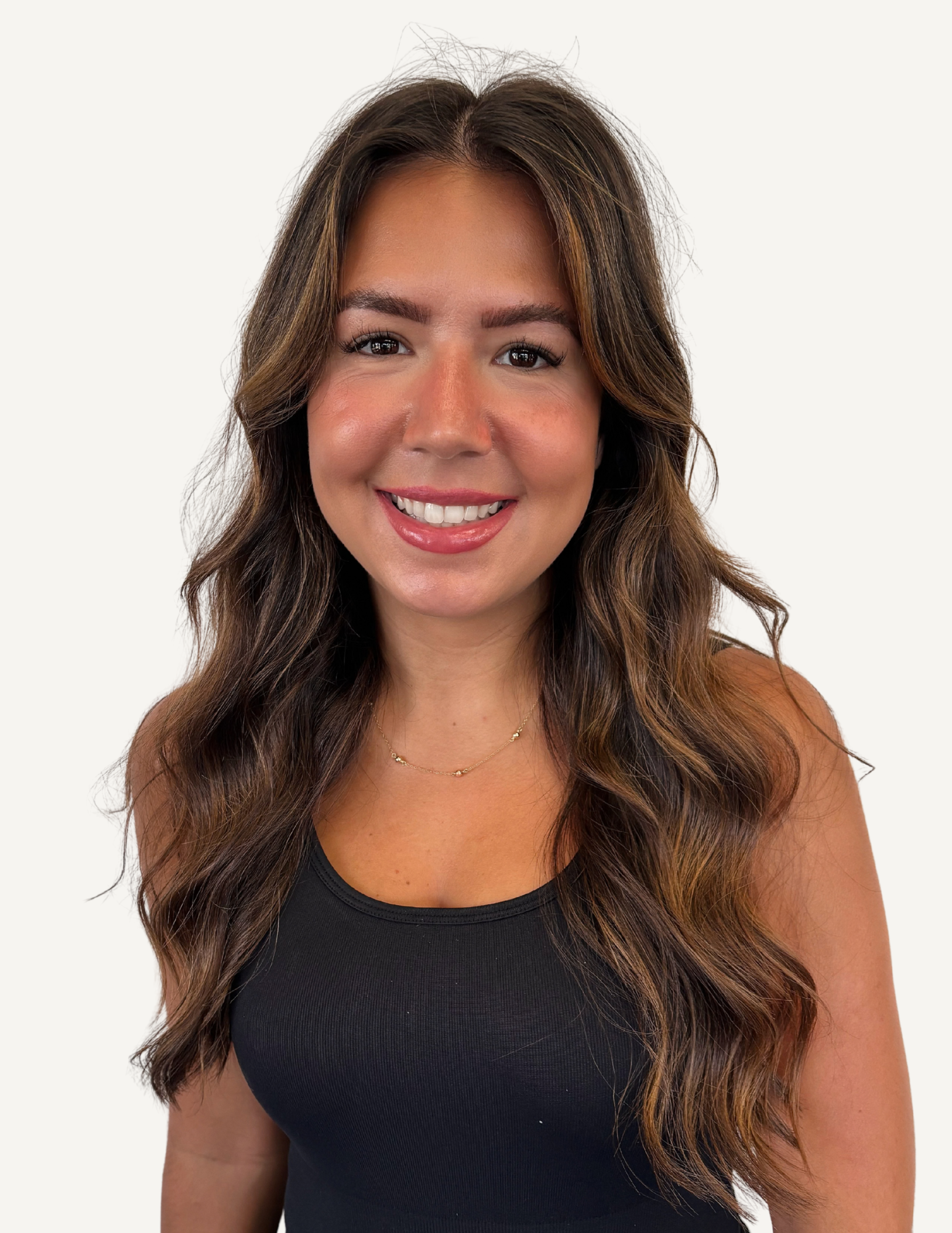 A woman with long wavy brown and blonde hair smiling indoors, wearing a black sleeveless top.