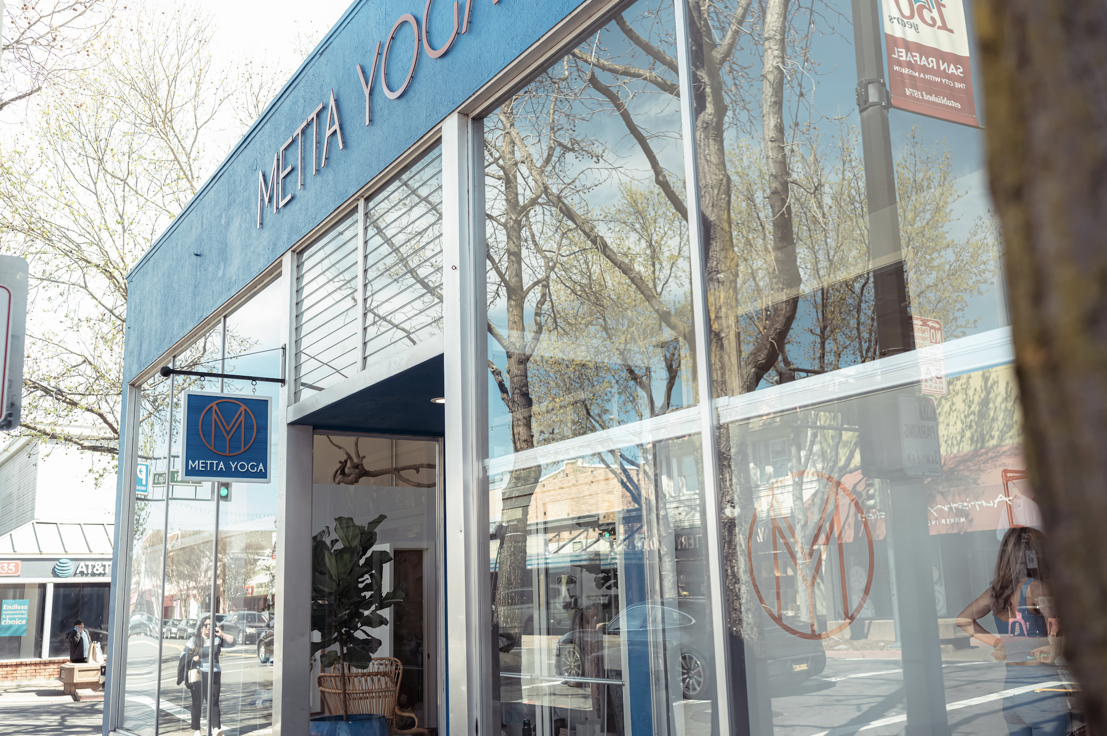 Exterior of a building with large glass windows reflecting trees and the sky, with a blue sign that reads 'Metta Yoga' hanging outside.