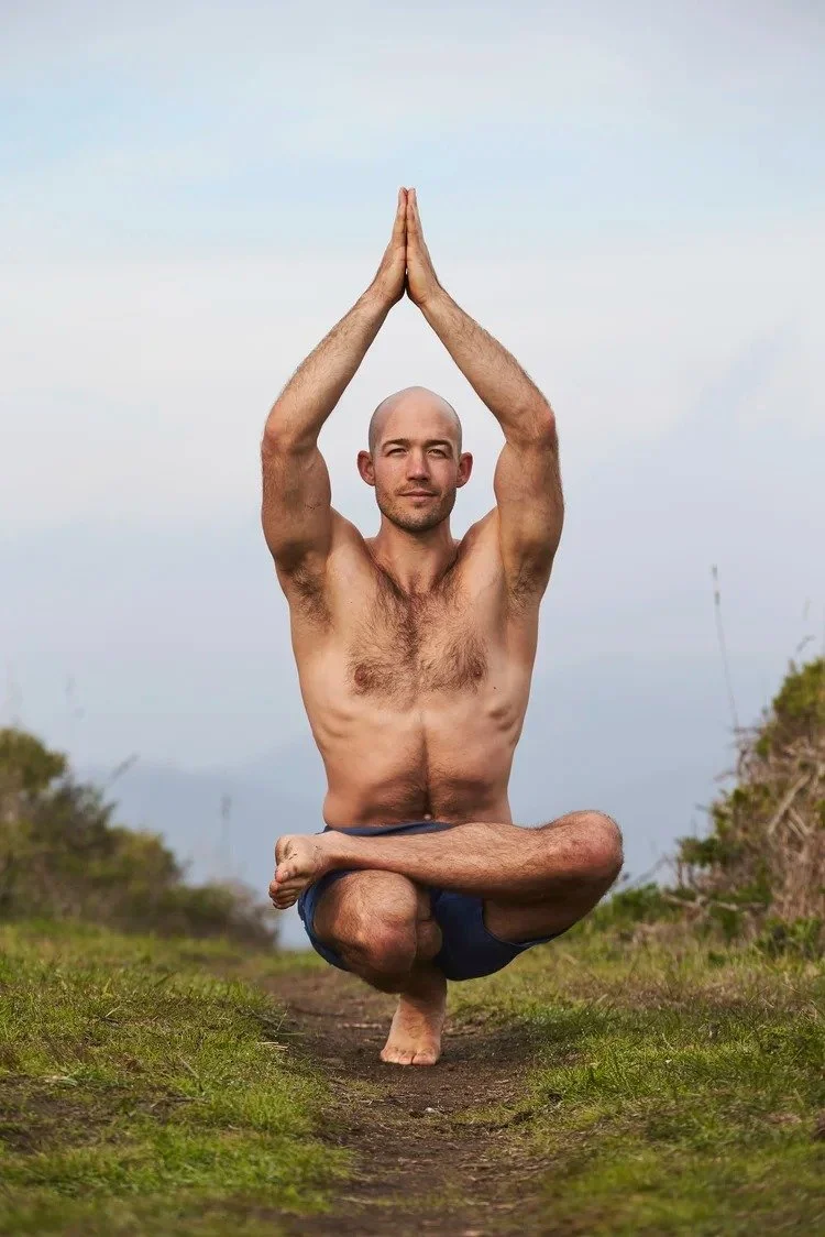 A shirtless man practicing yoga outdoors, balancing on one foot with his hands pressed together above his head in a prayer position, sitting on a grassy path with a cloudy sky in the background.