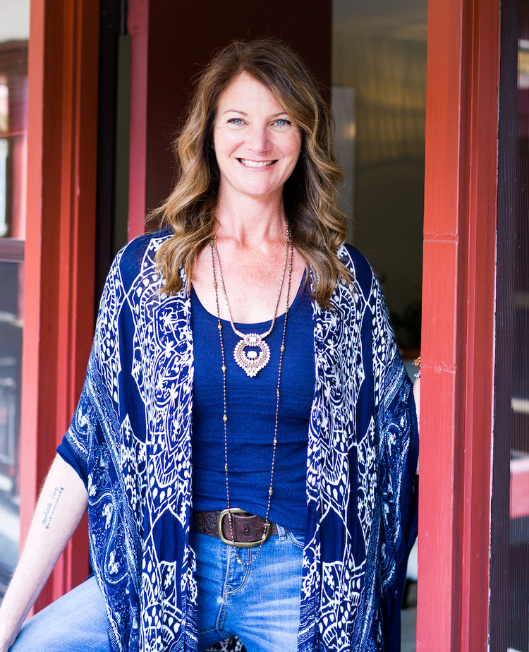 A woman with long wavy brown hair smiling, wearing a blue patterned kimono over a blue top, blue jeans, and layered necklaces, standing near a red door and window.