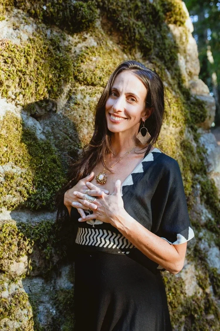A woman with long dark hair, wearing a black and white dress and jewelry, standing outdoors near a moss-covered rock wall, smiling at the camera.