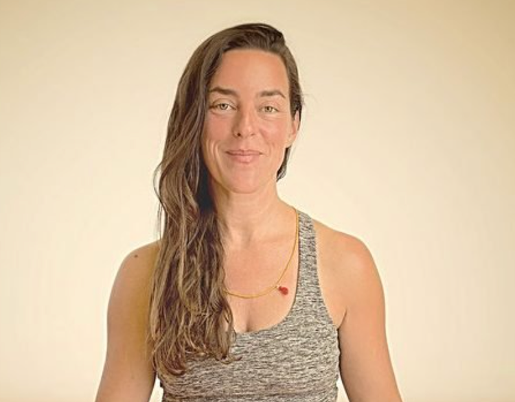 A woman with long wavy brown hair wearing a sleeveless gray top and a gold necklace with a small red pendant, smiling slightly against a plain light-colored background.