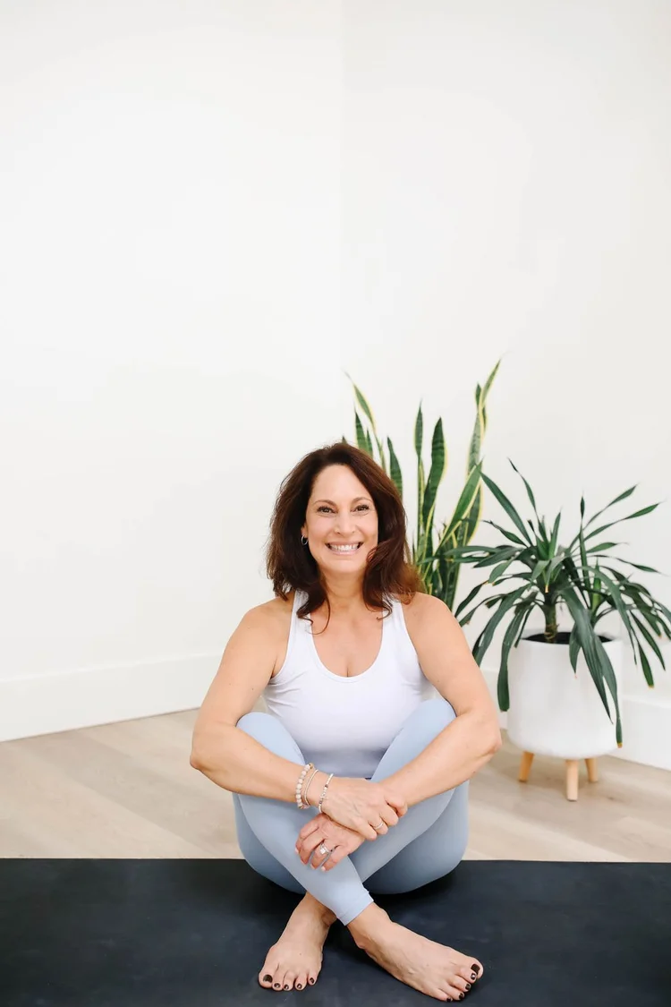Smiling woman practicing yoga seated on a mat with hands clasped around her knees, indoor setting with plants in white pots in the background.