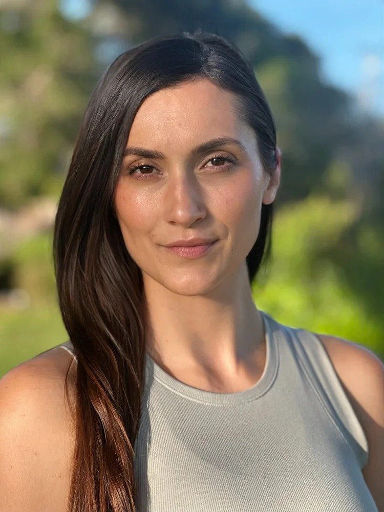 A woman with long dark hair outdoors, smiling softly, wearing a sleeveless gray top with a blurred green and blue background.
