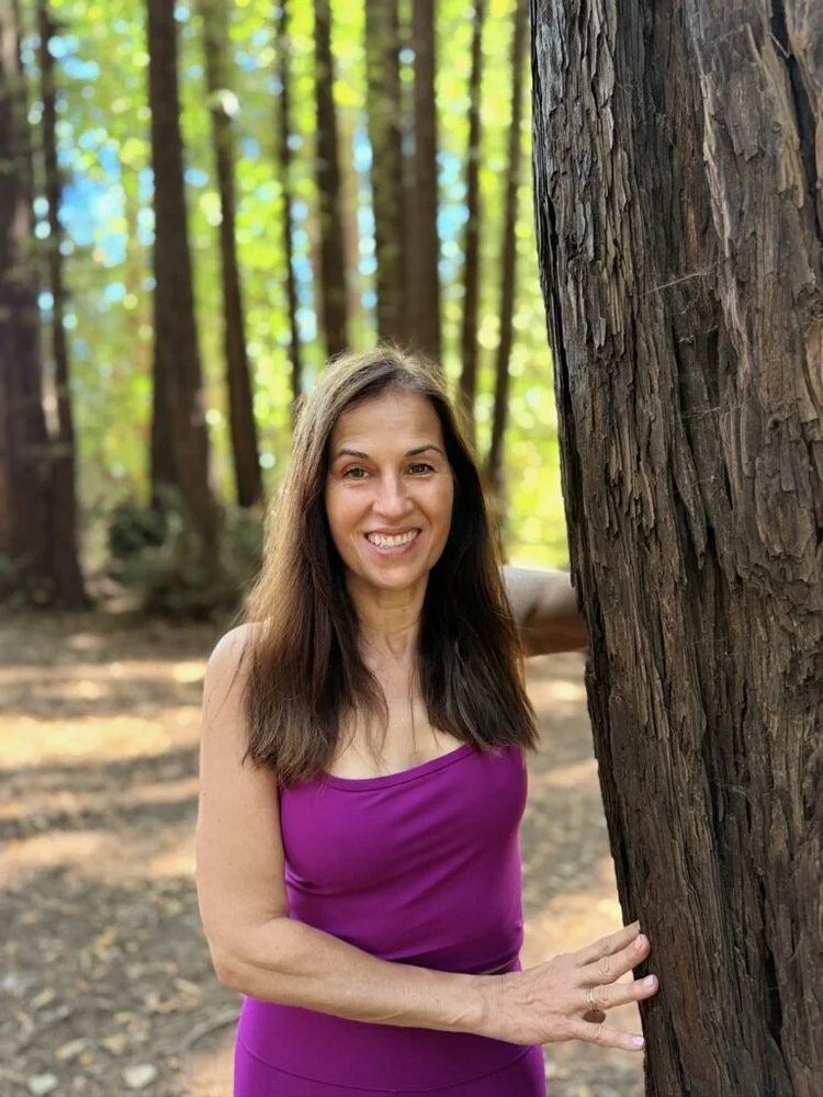 A woman with long brown hair wearing a purple tank top stands outdoors in a forest, smiling and touching a large tree trunk.