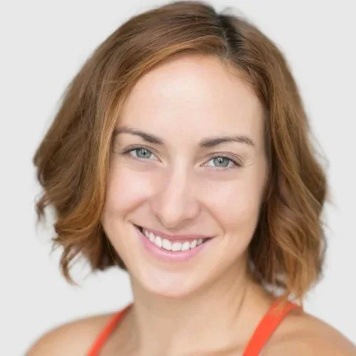 A young woman with shoulder-length wavy red hair, blue eyes, and a big smile, wearing an orange tank top against a plain white background.