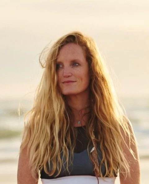 Woman with long blonde hair standing outdoors near a body of water at sunset.