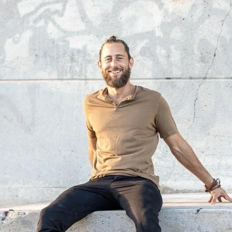 A man with a beard and long hair tied back, wearing a camel-colored t-shirt and black pants, sitting on a concrete ledge against a plain concrete wall, smiling at the camera.