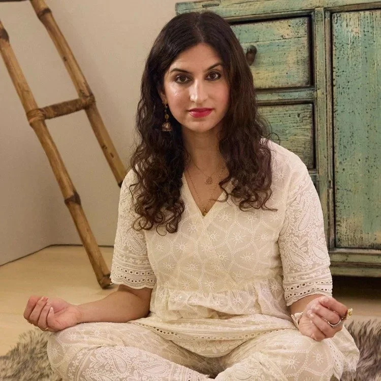 A woman with dark curly hair sitting cross-legged on a furry rug, wearing a cream-colored embroidered romper, with wooden furniture and a ladder in the background.