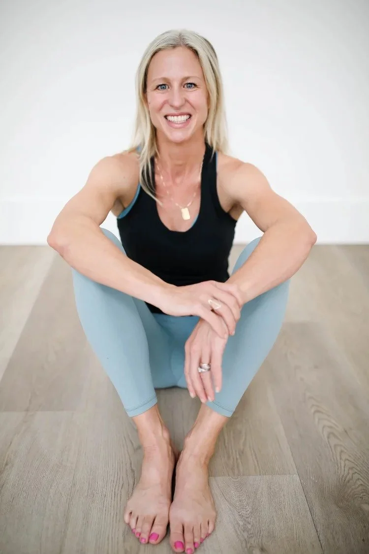 A smiling middle-aged woman with blonde hair, wearing a black tank top and light blue leggings, sitting on the floor with her legs crossed and hands resting on her knees.