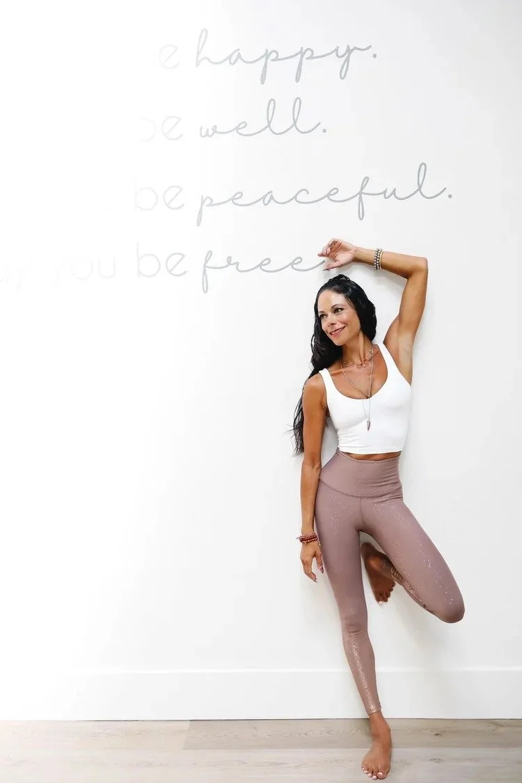 A woman with long dark hair poses against a white wall, smiling, with one hand resting on her head. She wears a white sleeveless crop top and light pink, sparkly leggings. Behind her, the wall has faint gray text that says, 'be happy, be well, be peaceful, be free.'