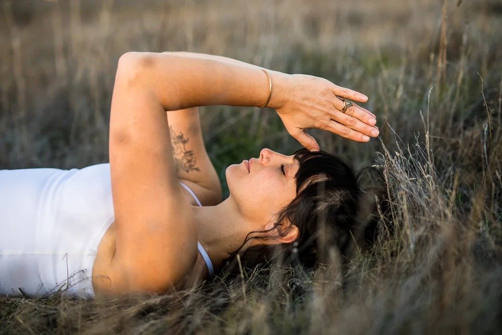 A woman lying on her back in tall grass with her eyes closed, raising one arm over her face in a serene outdoor setting.