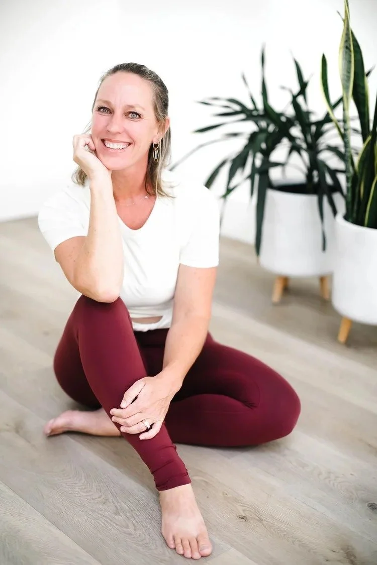 A woman with gray hair sitting cross-legged on a hardwood floor, smiling, wearing a white t-shirt and maroon leggings, with a large potted plant in the background.