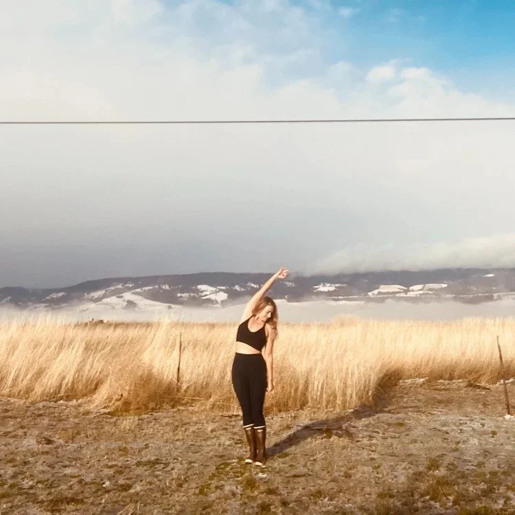 A woman in black clothing standing on a gravel path in a field of tall golden grass, stretching her arm upwards, with a backdrop of mountains and a partly cloudy sky.