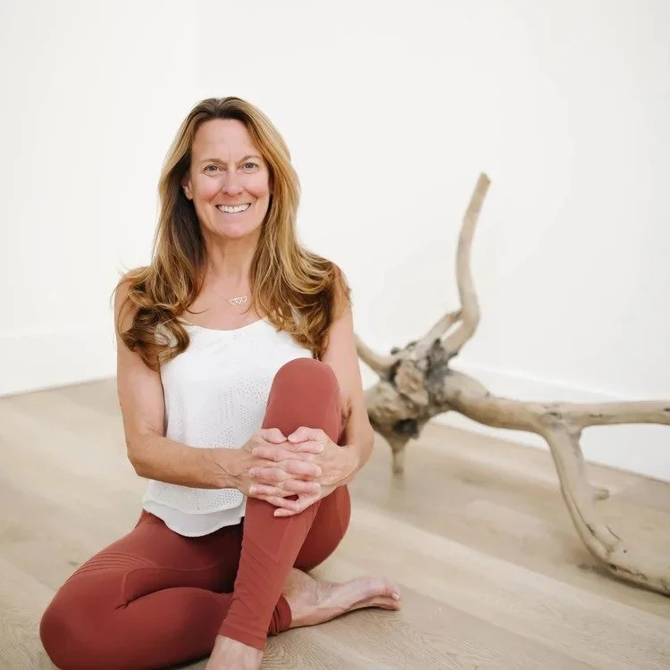 A woman with long, wavy, light brown hair sitting on a wooden floor in a yoga pose, smiling, with a white wall and a piece of driftwood in the background.