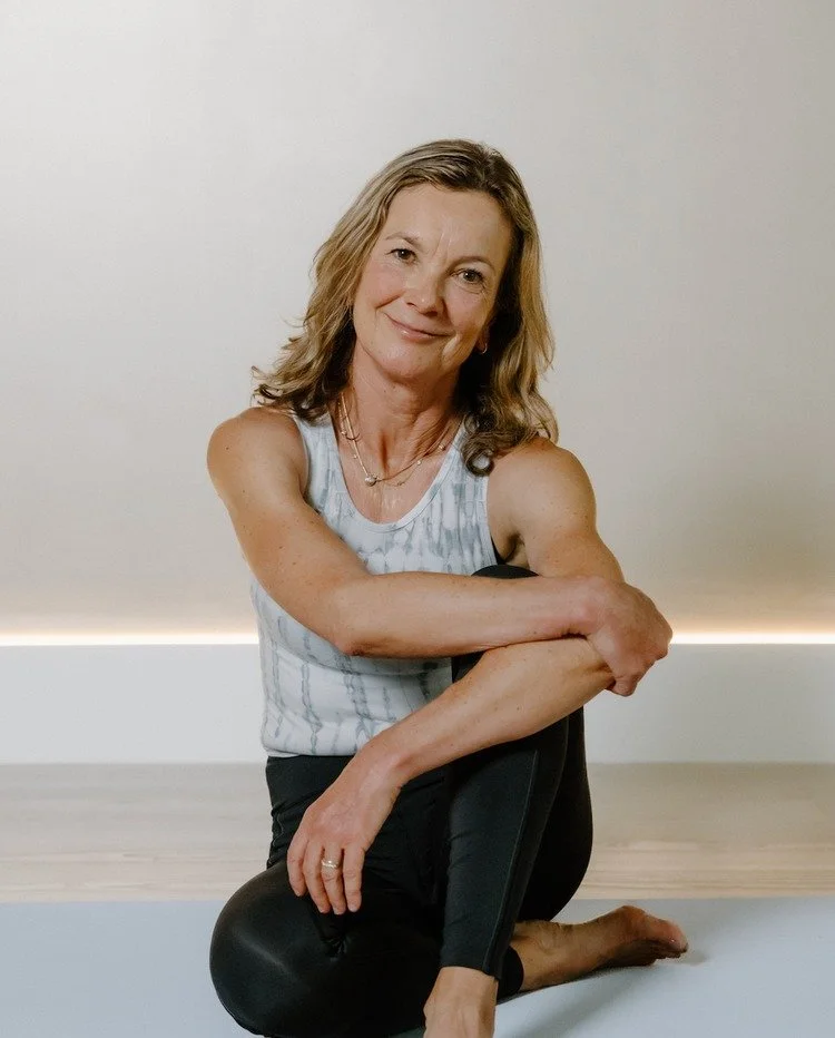 A woman sitting on the floor in a yoga pose, hugging her knees, smiling at the camera, in a room with light-colored walls and floor.