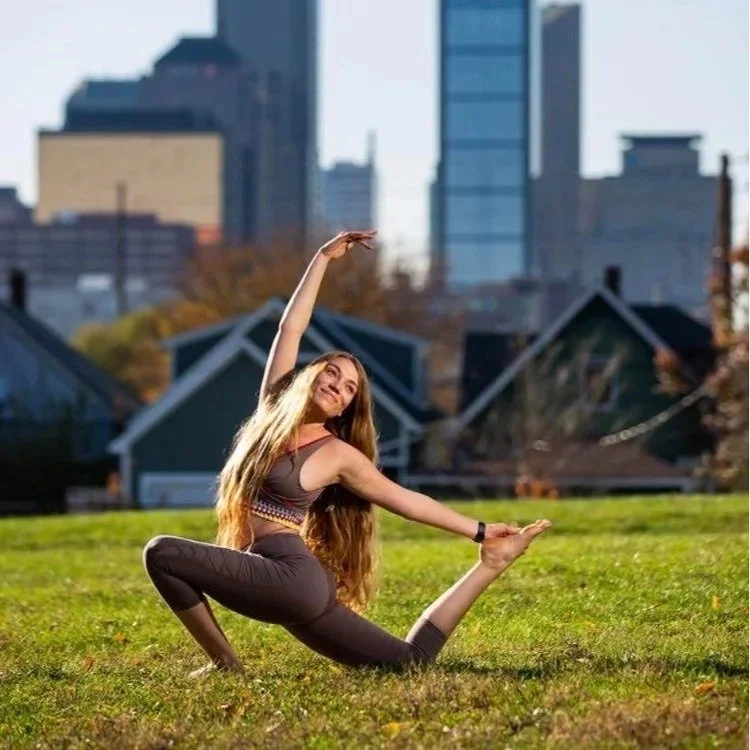 A woman practicing yoga outdoors in an urban park during daytime, with city buildings in the background.