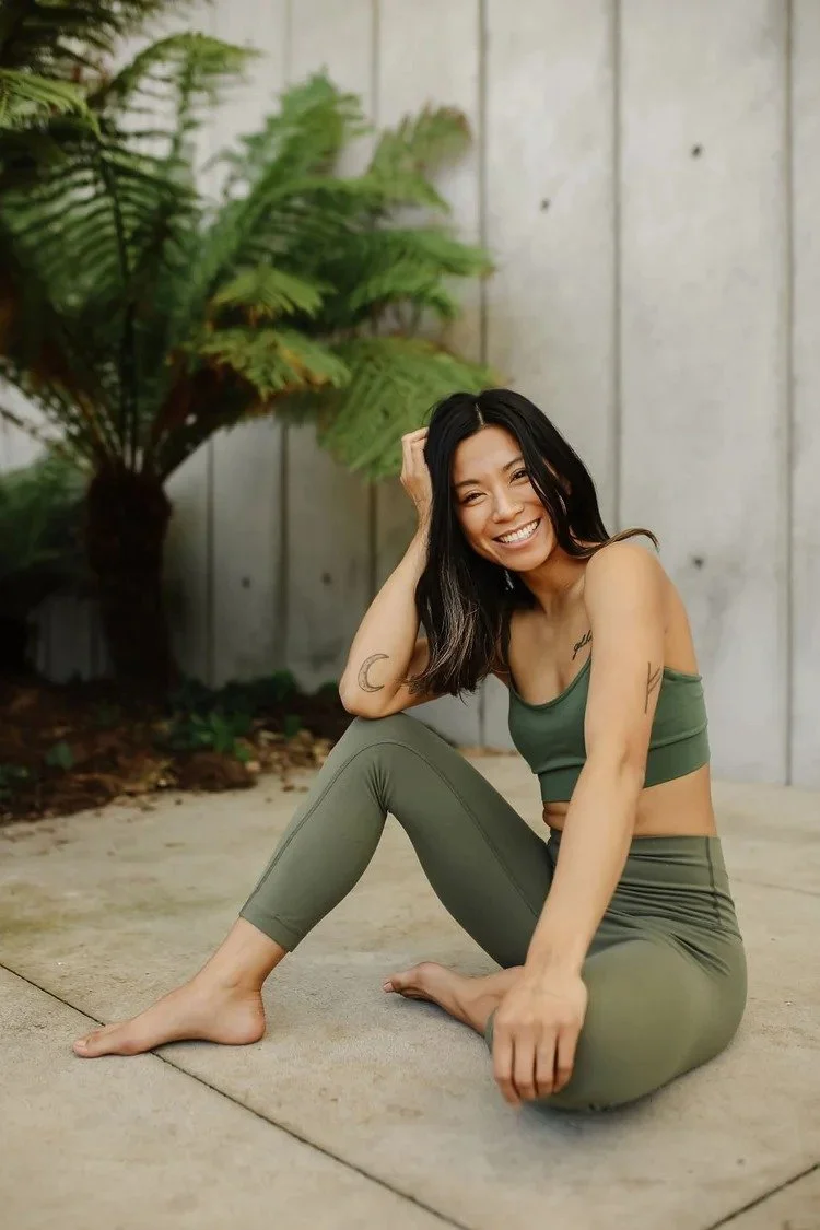 A smiling woman with dark hair, sitting on the ground outside, wearing an olive green sports bra and matching leggings, with a background of a wooden fence and large greenery.