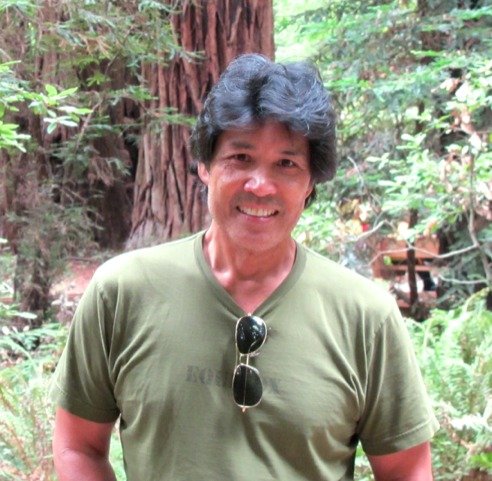 A man with dark hair and a broad smile, wearing a green t-shirt and sunglasses hanging from the collar, standing outdoors in a forested area with trees and foliage in the background.