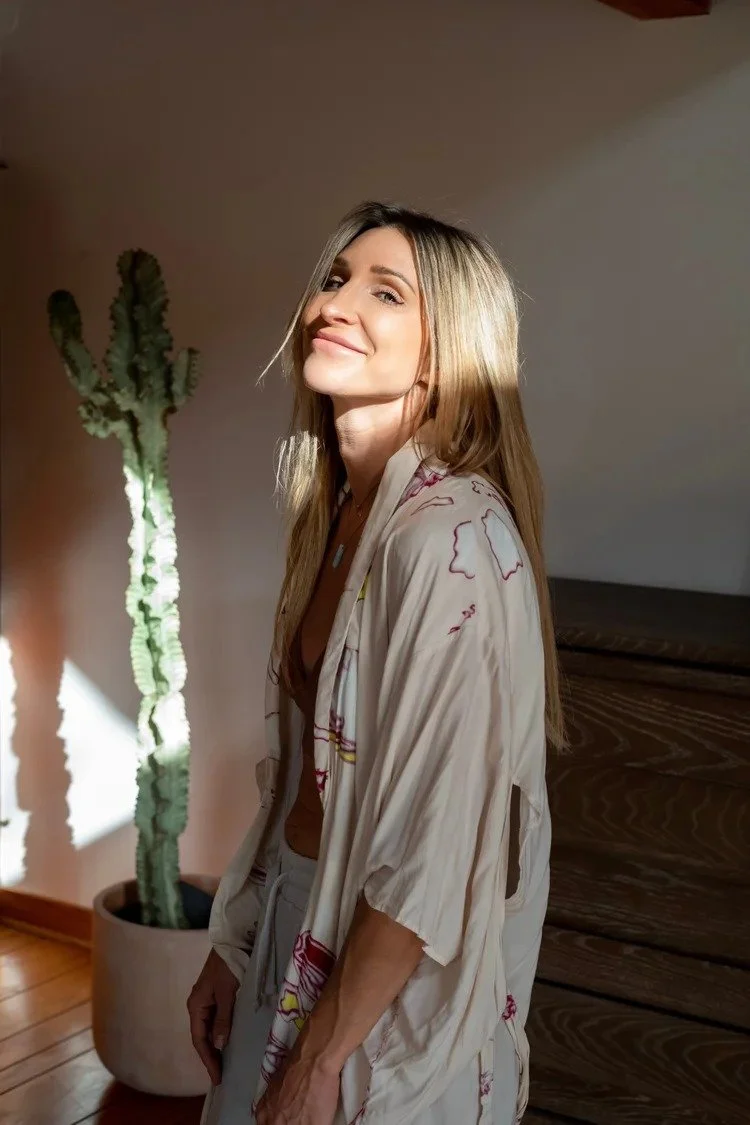 A woman with long blonde hair smiling and standing indoors next to a tall cactus plant, wearing a light-colored pajama top.
