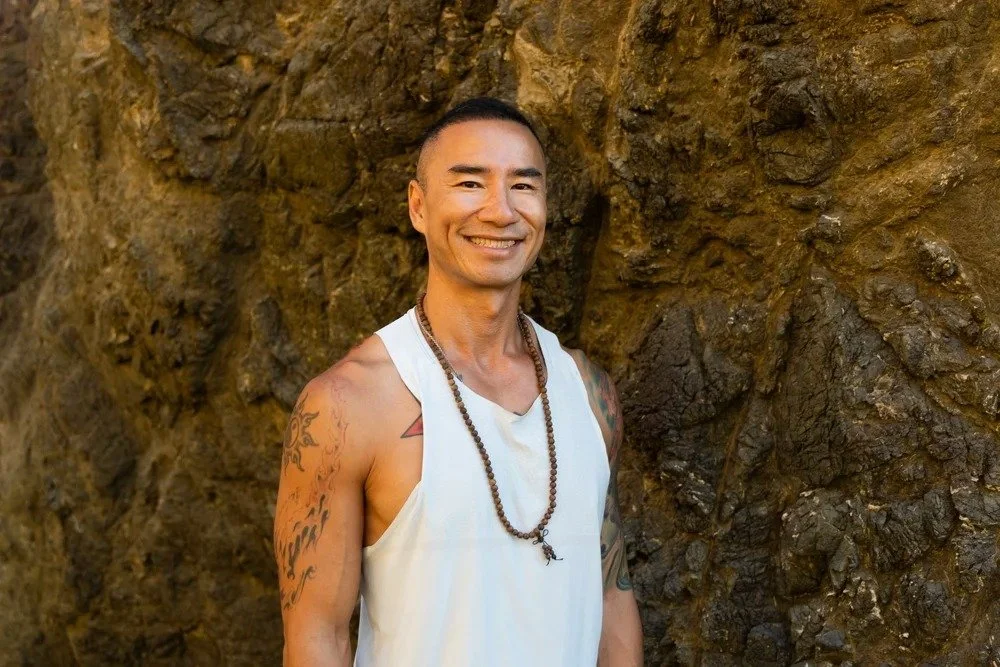 Smiling man wearing a white sleeveless shirt and a beaded necklace standing in front of a rocky background.