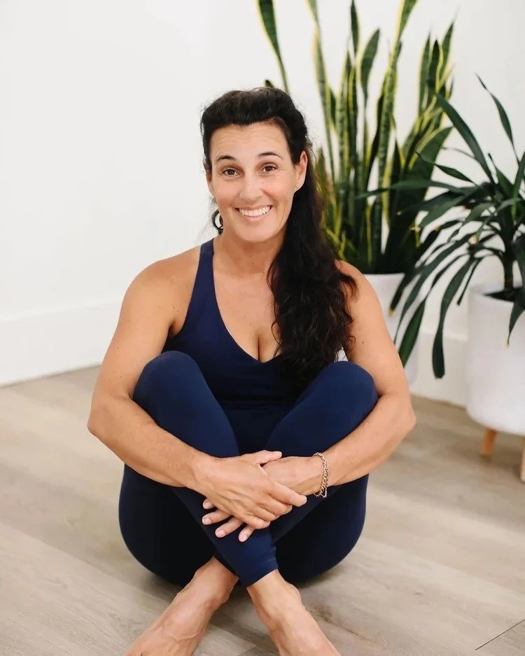 A woman with long dark hair in a ponytail, wearing a navy tank top and matching leggings, sitting cross-legged on the floor with a big smile, in a room with wooden flooring and potted plants.