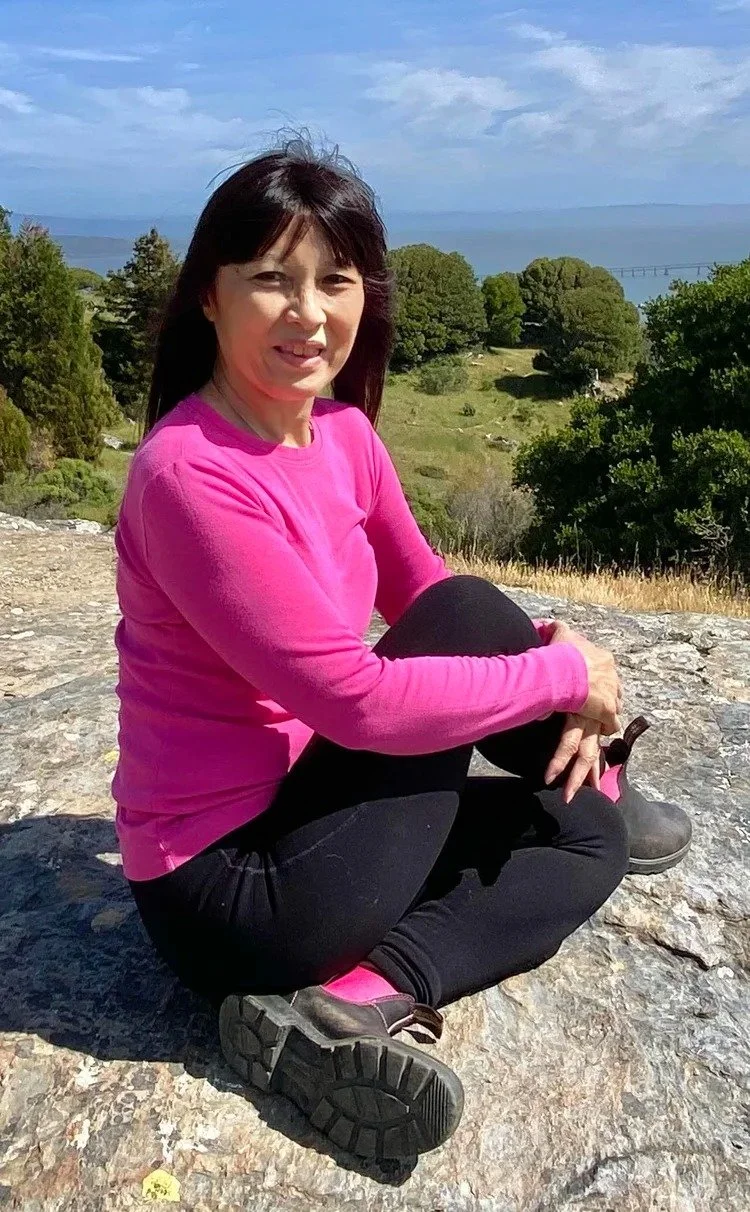 Woman sitting cross-legged on rocky ground outdoors, wearing a pink long-sleeve shirt and black pants, with trees, a grassy landscape, and a bridge in the background under a blue sky with scattered clouds.