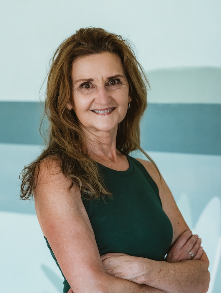 A smiling middle-aged woman with long, wavy, reddish-brown hair, wearing a sleeveless dark green top, standing with arms crossed against a light, abstract background.