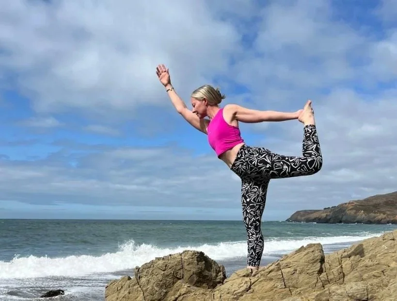 A woman practicing yoga on rocks at the beach, balancing on one leg and holding her other foot behind her back, with the ocean and cloudy sky in the background.