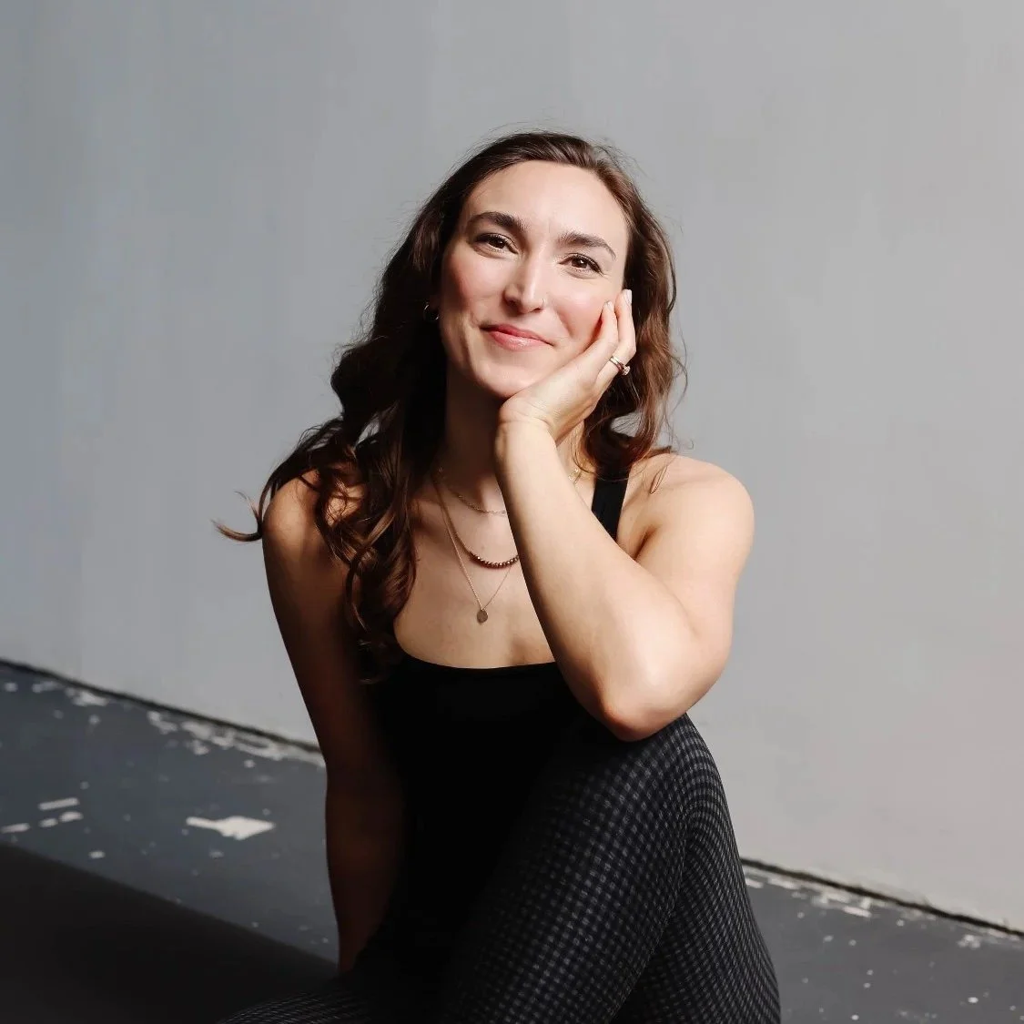 A young woman with long brown hair and a black tank top sitting on the floor, smiling softly with her hand resting on her face, against a plain gray background.