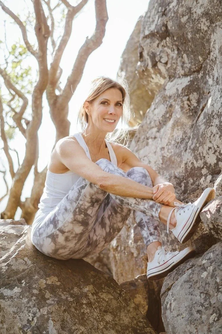 A woman smiling and sitting on rocks outdoors, wearing athletic clothing and sneakers, with trees in the background.