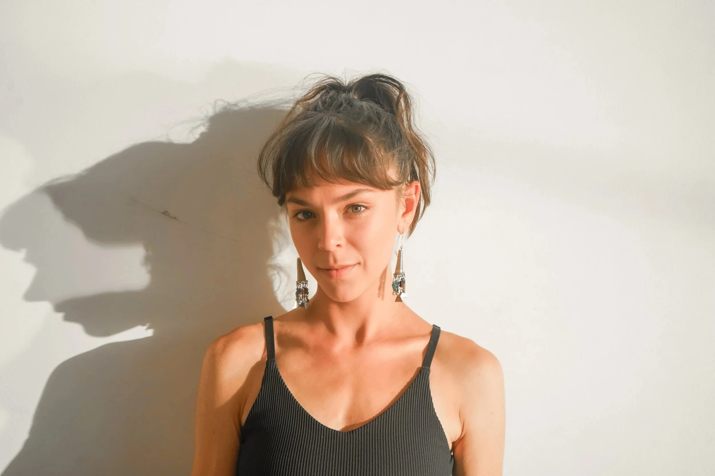 A young woman with brown hair tied in a high ponytail, wearing a black sleeveless top and decorative earrings, stands against a plain light-colored wall with a shadow cast to her left.