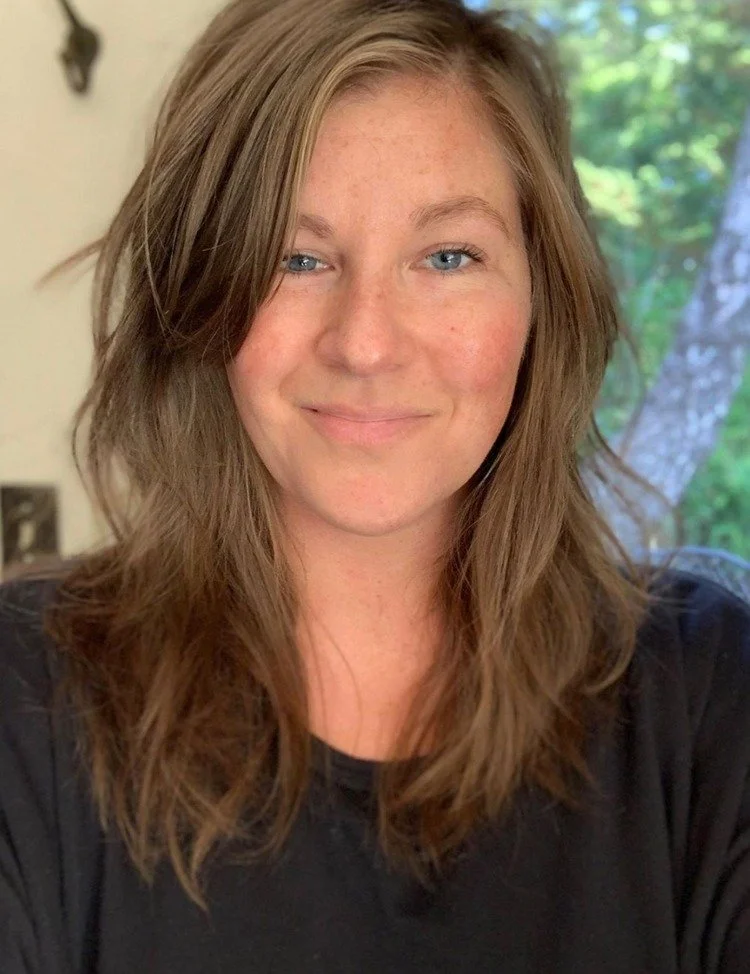 A woman with shoulder-length wavy brown hair, blue eyes, and fair skin, smiling softly, wearing a black top, with a window and greenery in the background.