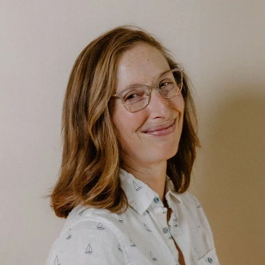 A young woman with shoulder-length red hair, wearing glasses and a white shirt with small sailboat prints, smiling and standing against a plain wall.