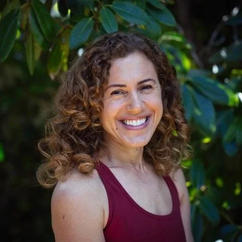A woman with curly hair smiling outdoors in front of green foliage, wearing a sleeveless maroon top.