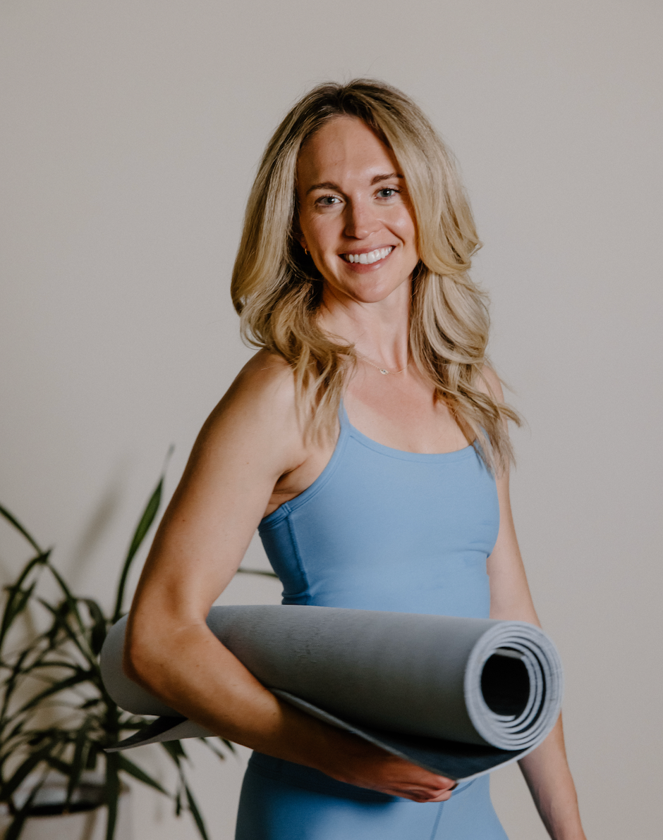 A woman smiling while holding a rolled yoga mat, wearing a blue workout top, with a plant in the background.