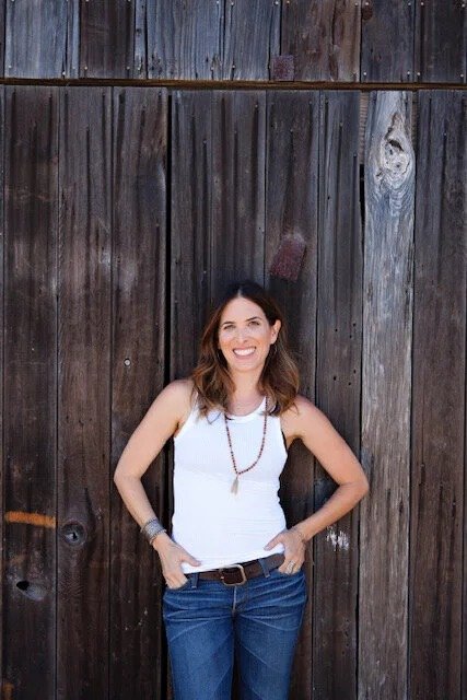 Woman smiling and standing in front of a wooden fence, wearing a white shirt, jeans, and a beaded necklace.