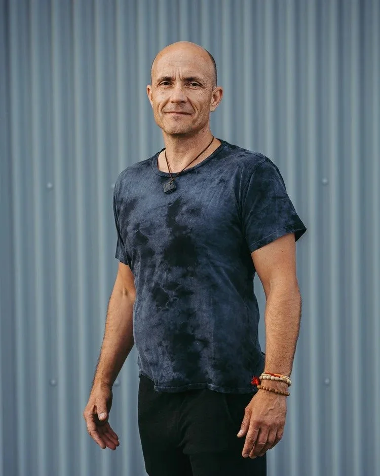 A middle-aged man with a shaved head and light skin, wearing a navy blue tie-dye T-shirt, black pants, and multiple bracelets, standing against a blue corrugated metal wall.