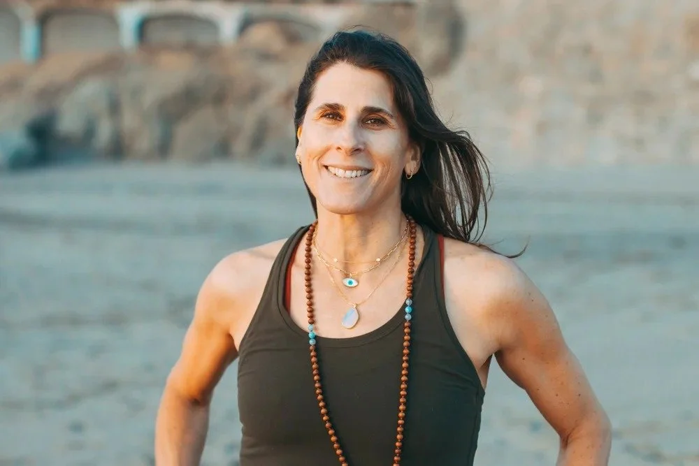 Smiling woman with long dark hair wearing a black tank top and layered necklaces on a beach.