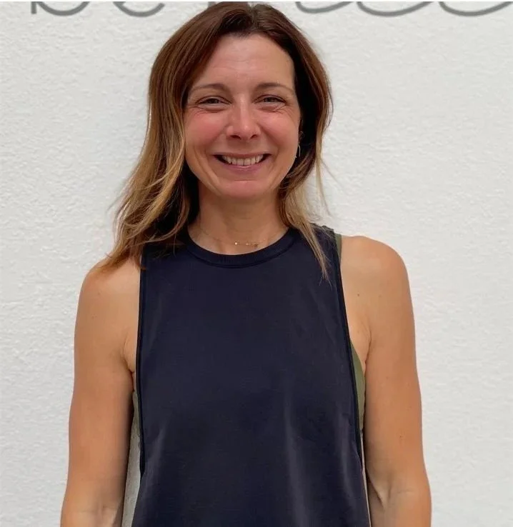 A smiling woman with shoulder-length brown hair, wearing a sleeveless black athletic top, standing against a plain white wall.