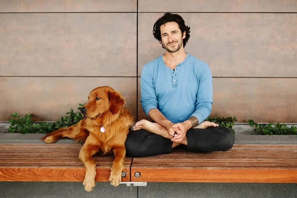 A man in a blue shirt sitting cross-legged on a wooden bench with a golden retriever dog beside him. They are outdoors against a tiled wall with some green plants behind them.
