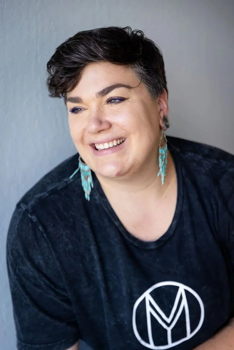 A smiling woman with short dark hair and earrings, wearing a black T-shirt with a white circular logo, against a plain gray background.