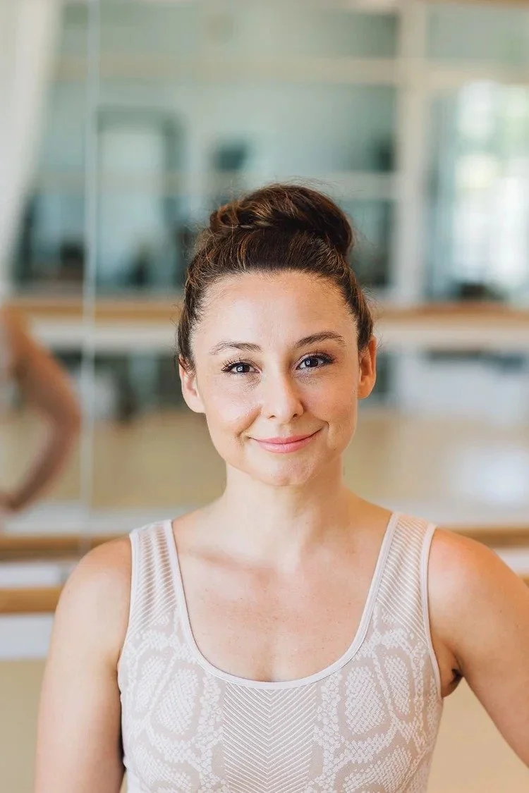 A young woman with dark hair in a bun, smiling and wearing a sleeveless top, standing in a dance studio with mirrors.