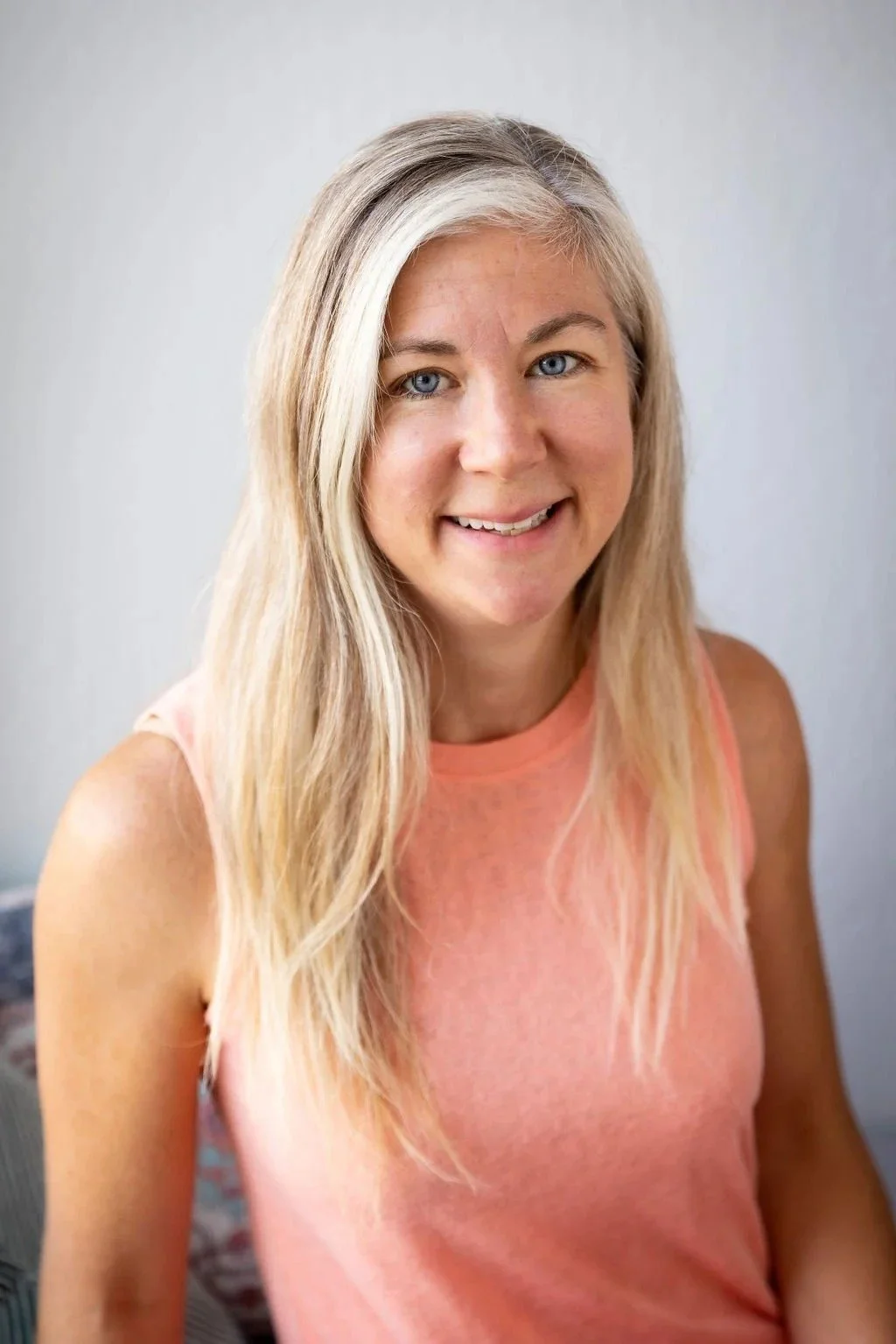A smiling woman with long blonde hair and blue eyes, wearing a sleeveless peach-colored top, standing indoors against a light-colored wall.