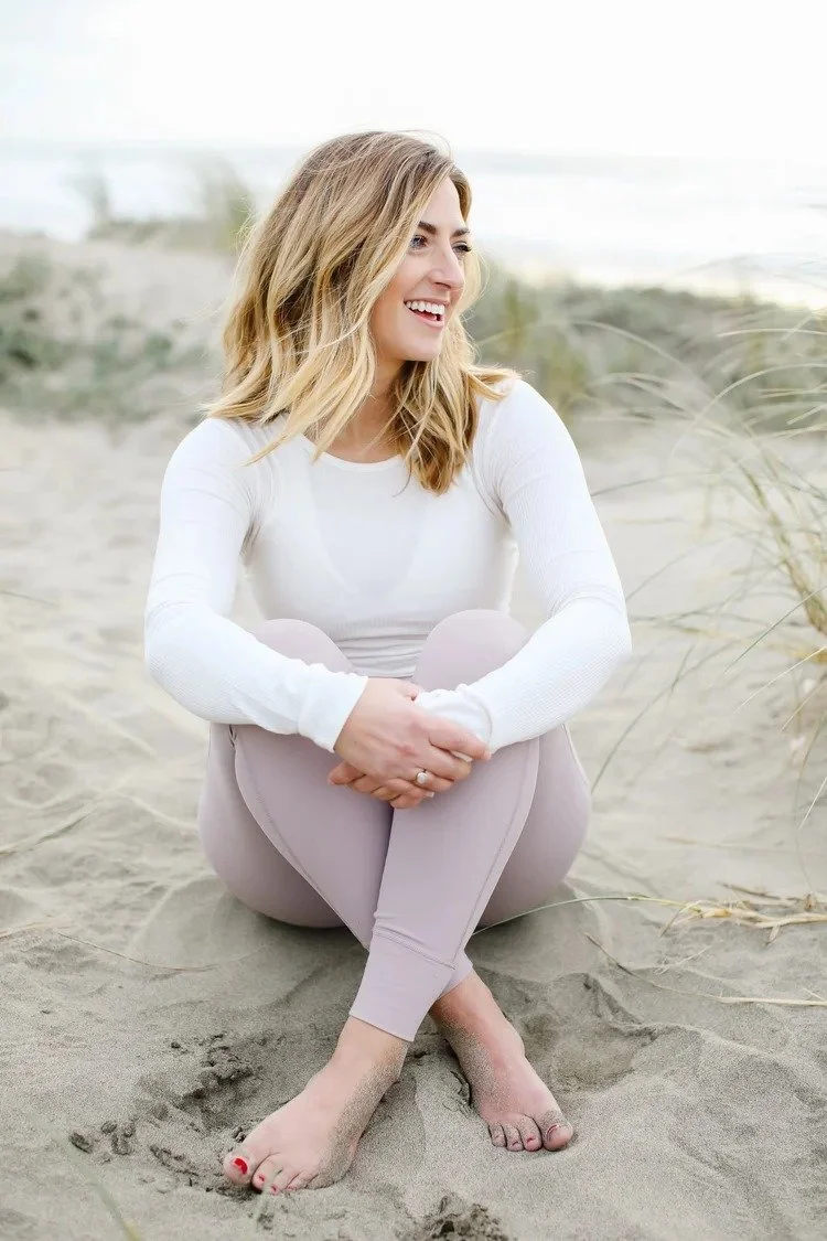 A woman with blonde hair sitting on the sand at the beach, smiling and looking away. She is wearing a white long-sleeve top and light-colored leggings. Sand dunes and grass are in the background.