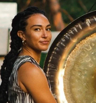 A woman with dark braided hair, gold earrings, wearing a black and white striped sleeveless top, standing next to a large metallic gong.