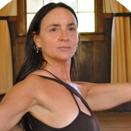 A middle-aged woman with long dark hair, wearing a black tank top, standing indoors with wooden walls and windows in the background.