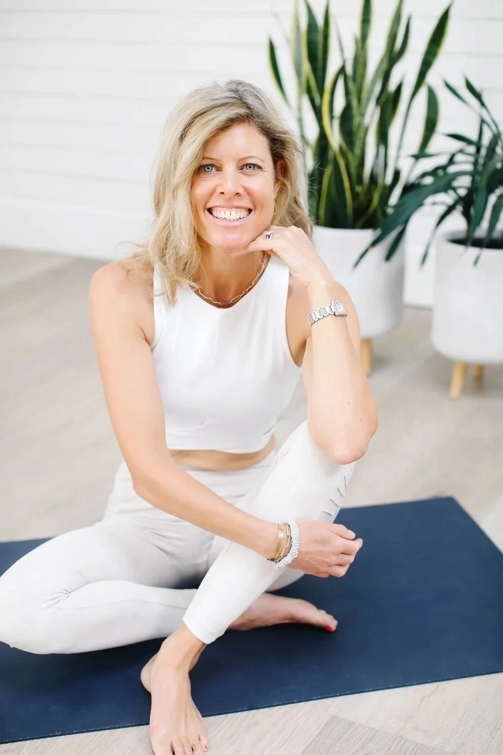A woman smiling while practicing yoga indoors, sitting on a black yoga mat with green plants in the background.