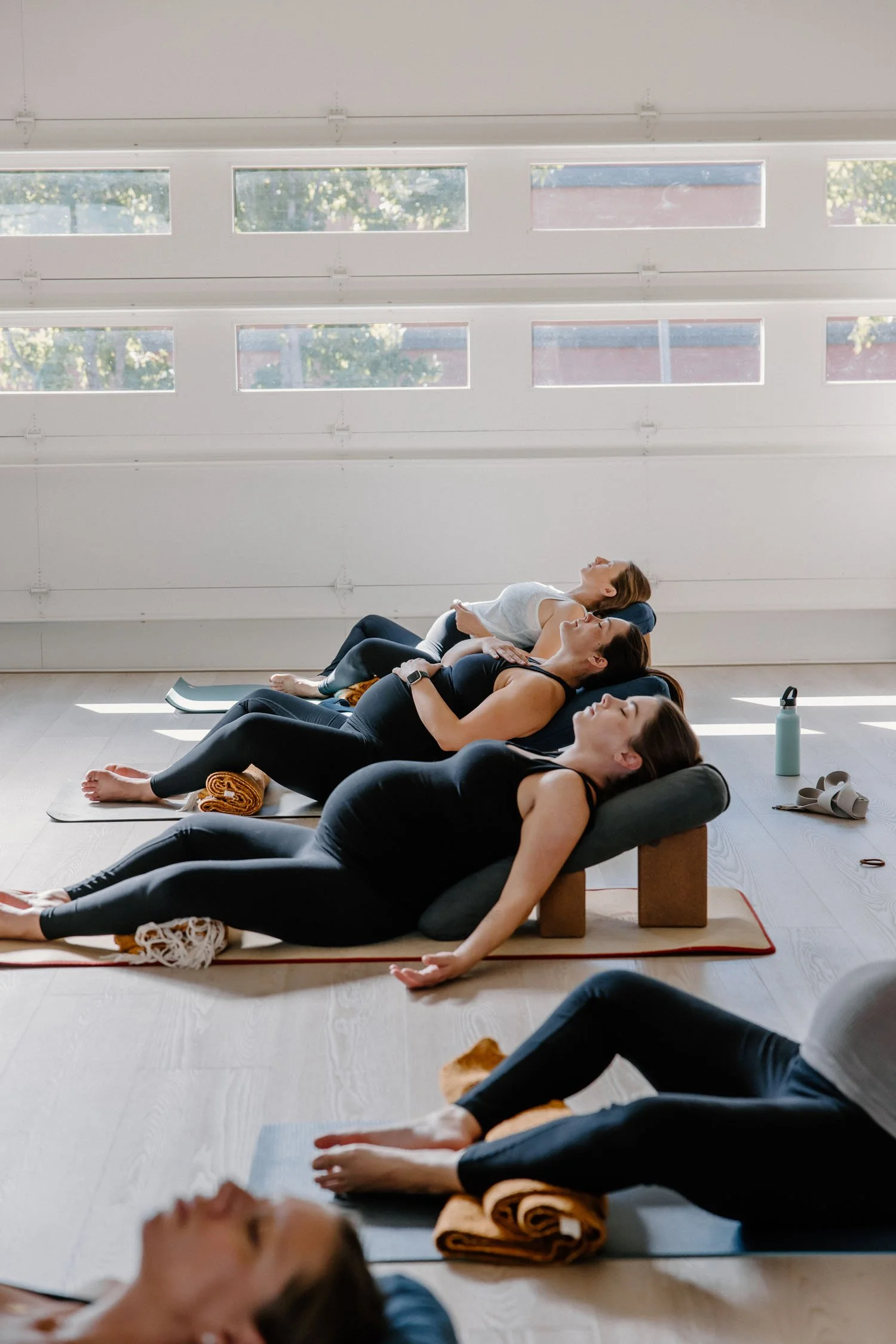 Women in a yoga class lying on their backs on yoga mats, relaxing with their eyes closed, in a bright studio with large windows.