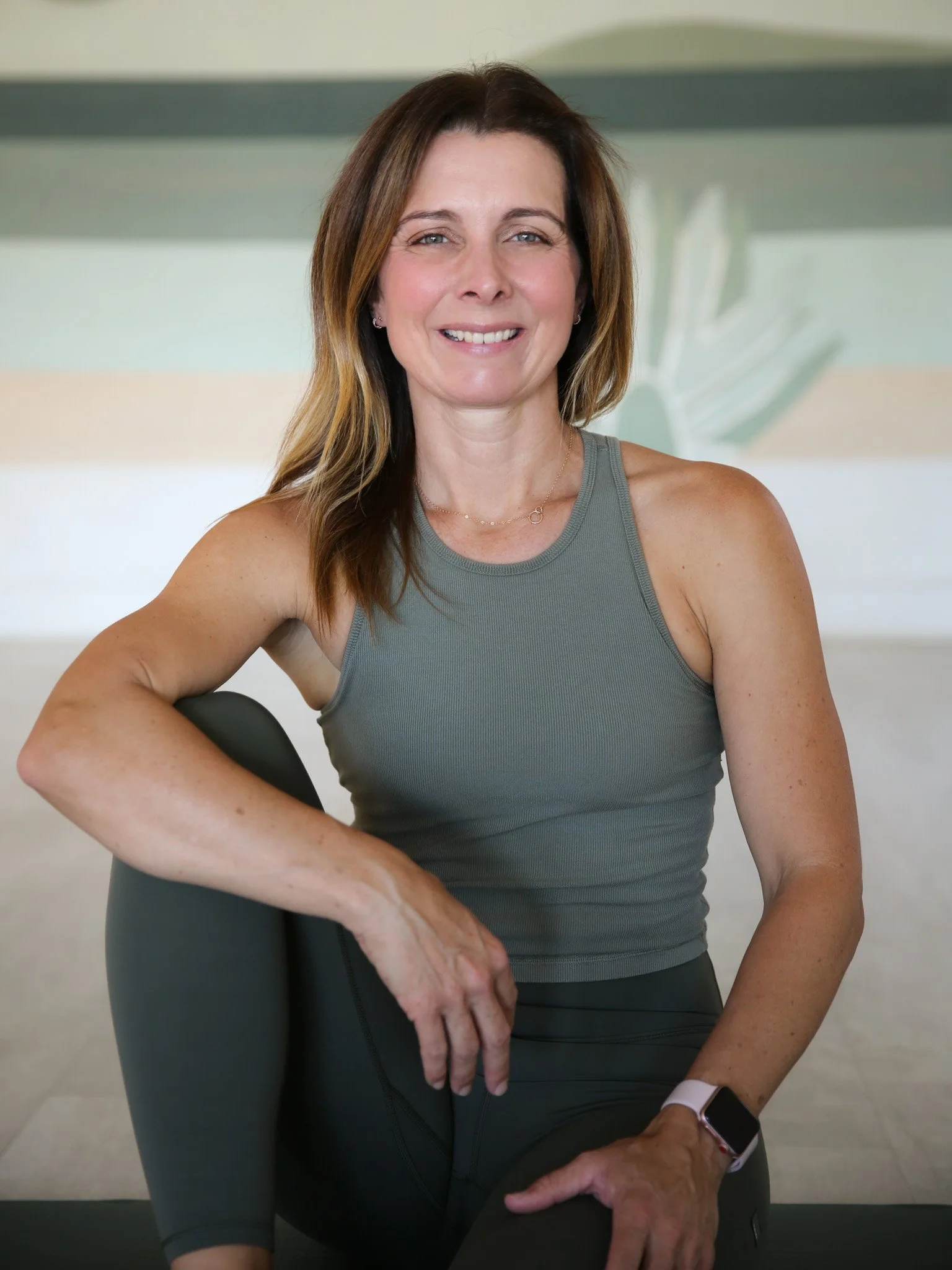 A smiling woman with shoulder-length brown hair, wearing a sleeveless black athletic top, standing against a plain white wall.